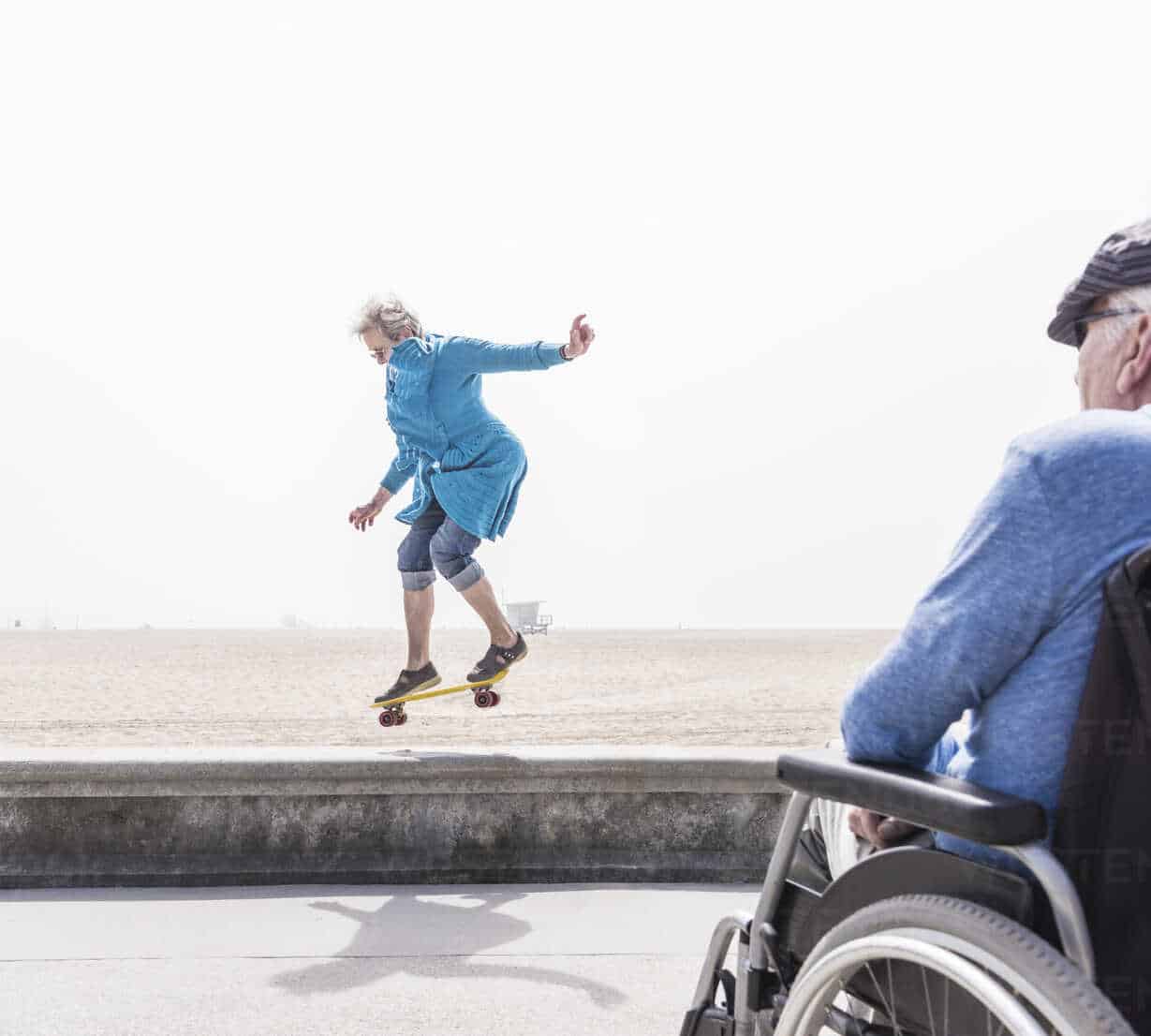Senior man in wheelchair watching wife doing skateboard trick at beach ...